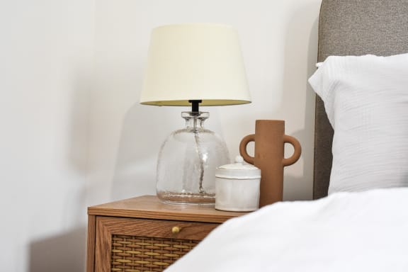 a nightstand with a lamp and a jar on it next to a bed
