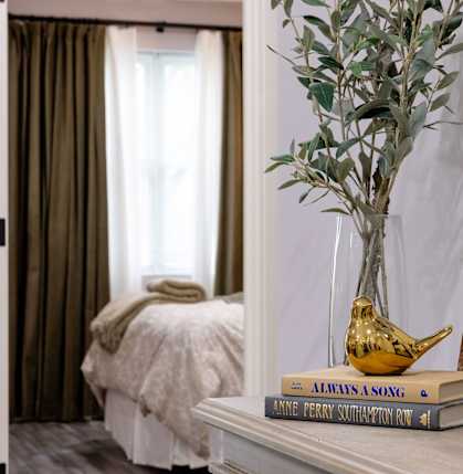 A room with a bed, a vase with a plant, a book, and a painting on the wall.