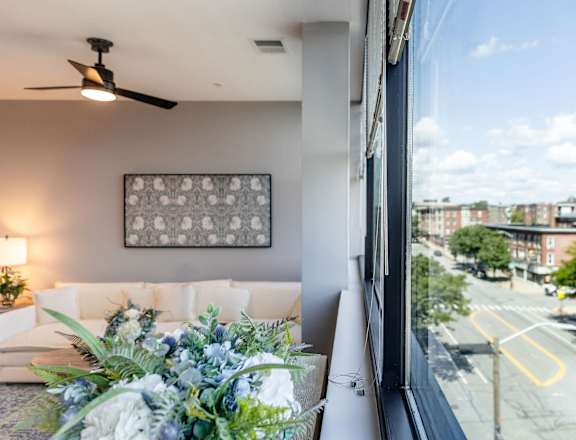 A living room with a couch, a fan, and a window overlooking a street.