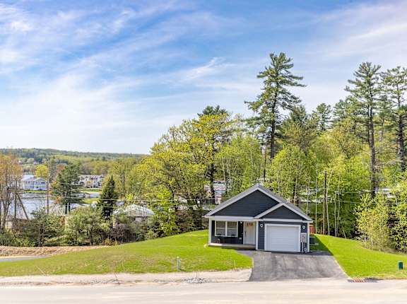 A house with a garage is situated on a grassy area with trees and a body of water in the background.
