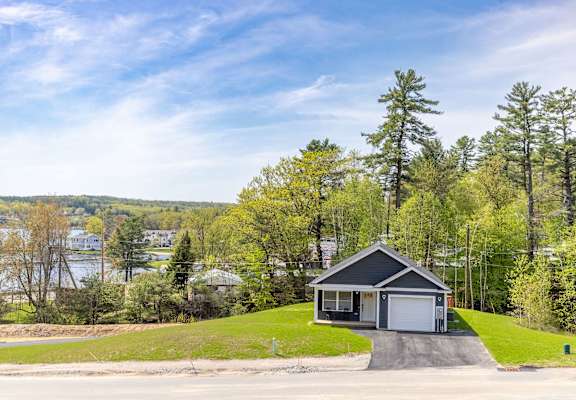 A house with a garage is situated on a grassy area with trees and a body of water in the background.