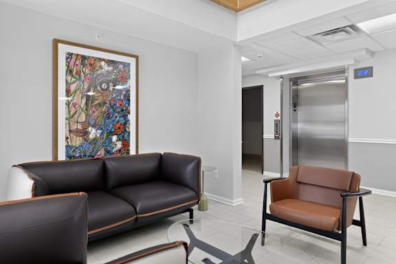 A brown leather chair sits in a waiting area with a painting on the wall and a silver elevator in the background.