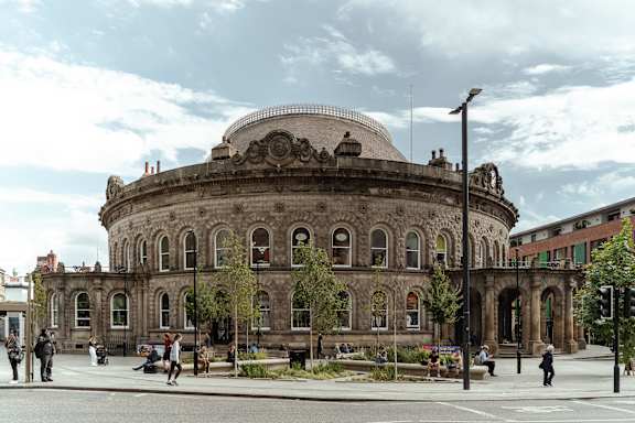 A large, old building with a dome on top is surrounded by a plaza with people walking and sitting.