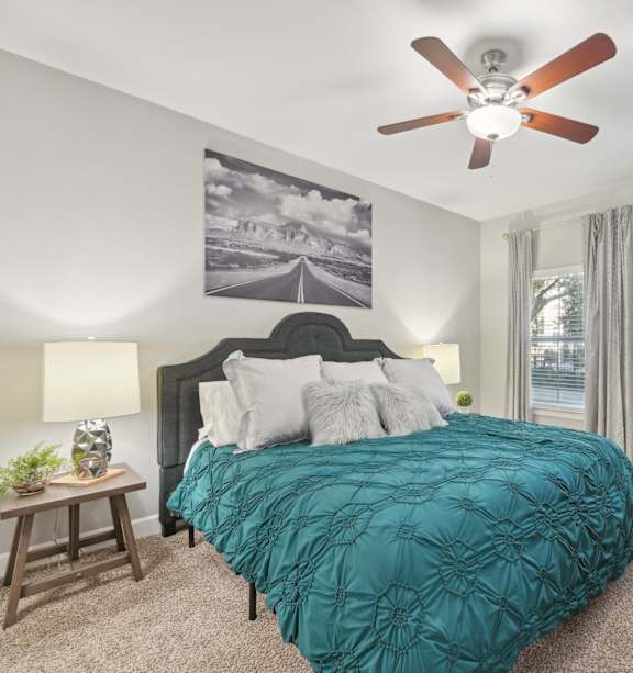 A bedroom with a bed, a ceiling fan, and a painting on the wall.