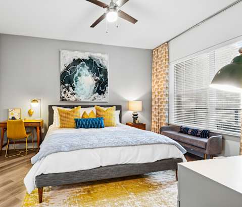 A bedroom with a bed, a chair, a painting, and a ceiling fan.