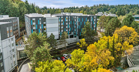A modern building with blue and white balconies is surrounded by trees with yellow leaves.