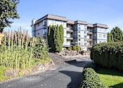 Thumbnail 8 of 8 - A large building with a grey and white exterior is surrounded by a well-manicured garden with a curved driveway leading up to it.