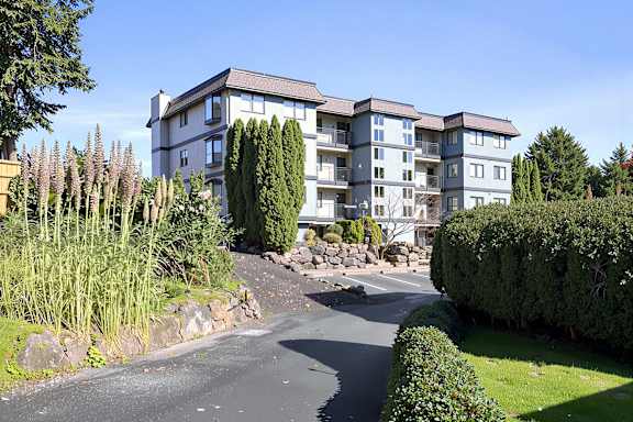 A large building with a grey and white exterior is surrounded by a well-manicured garden with a curved driveway leading up to it.