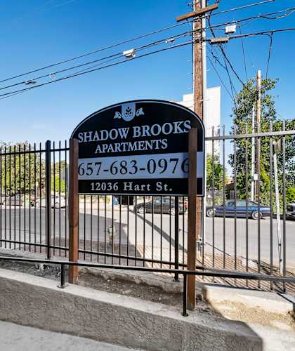 A sign for Shadowbrooks Apartments is displayed in front of a fence.