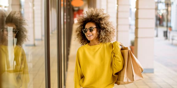 A woman with a yellow sweater and white pants is holding a brown paper bag while standing in front of a glass door.