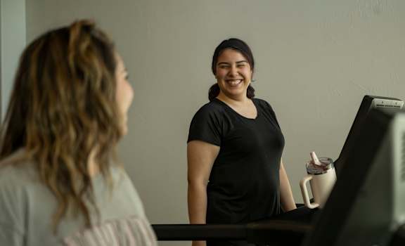 A woman in a black top is walking on a treadmill while holding a drink.
