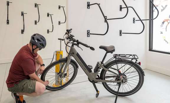 A man in a red shirt and grey shorts is fixing a bike in a room with a white wall and a black bike rack.