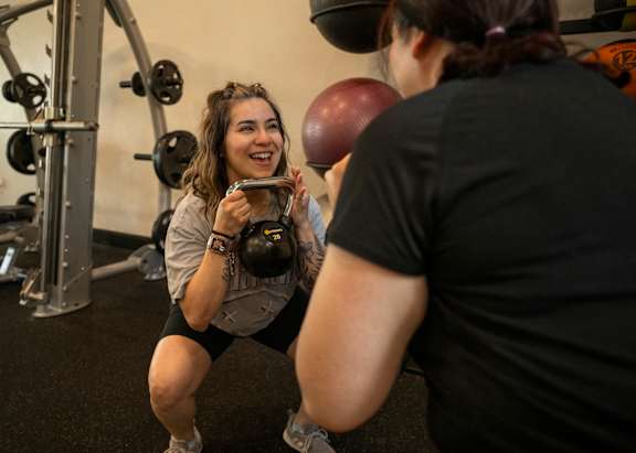 A woman is squatting in a gym holding a kettlebell while another woman stands behind her holding a medicine ball.