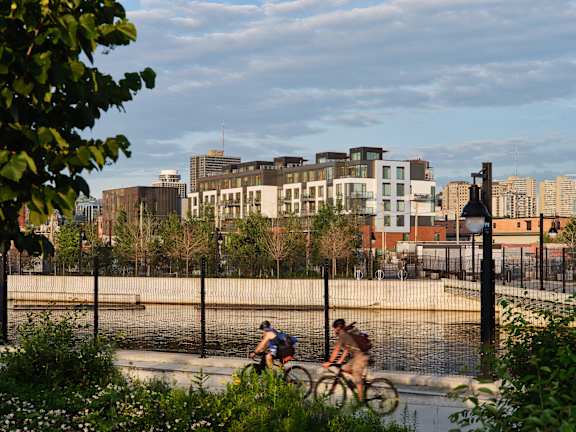 Two people riding bicycles on a path near a body of water with buildings in the background.