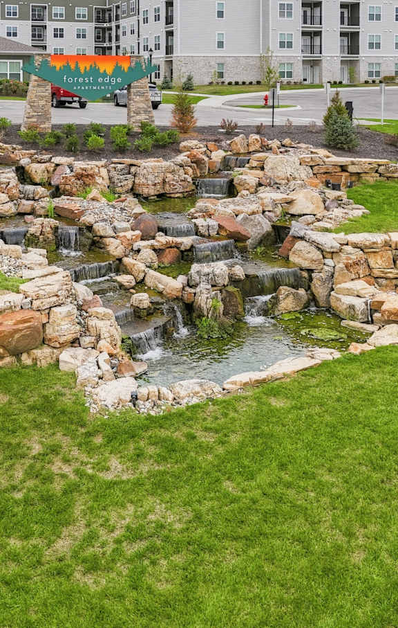 A small waterfall in a grassy area with a building in the background.