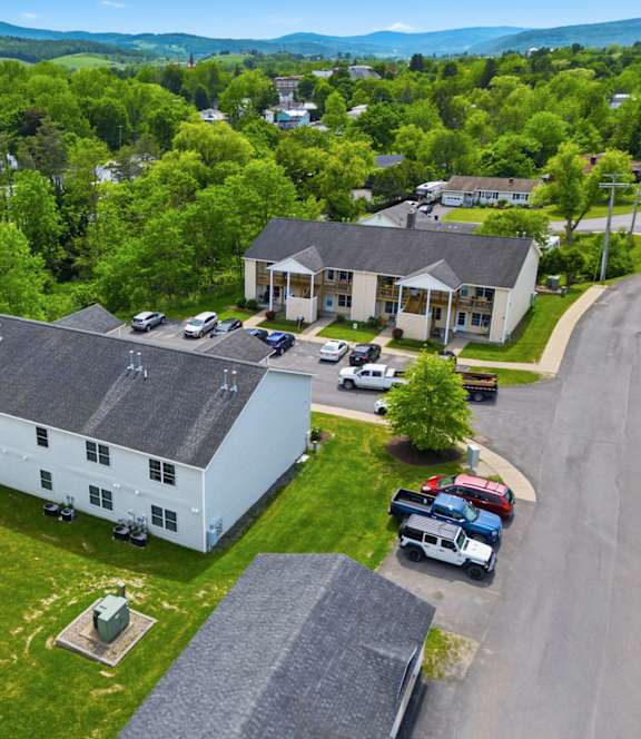 A white house with a black roof is surrounded by greenery and a parking lot.