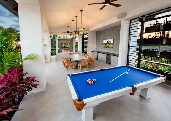 A pool table with a blue cloth and a white cue is in the center of a room with a ceiling fan and a television.