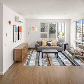 A living room with a grey couch, a coffee table, a chair, and a large window with a view of the street.