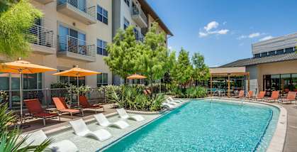 A large swimming pool surrounded by lounge chairs and umbrellas in front of a modern building.