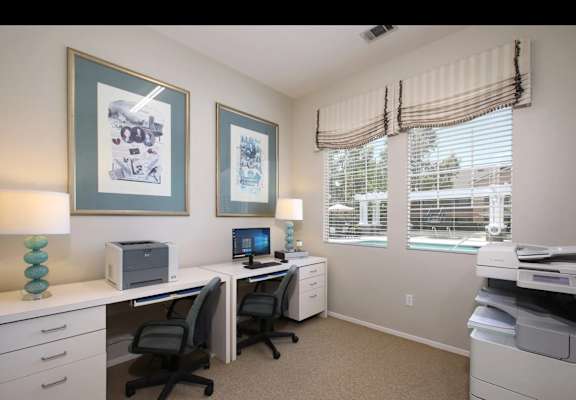 A well-lit office with two desks and chairs, a printer, and two framed pictures on the wall.