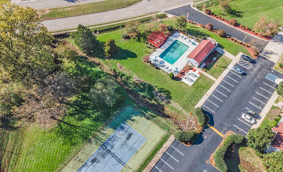 A tennis court is surrounded by a parking lot and a grassy area.
