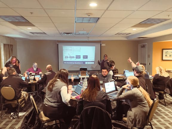 a group of people sitting around a table with laptops in front of a projector screen