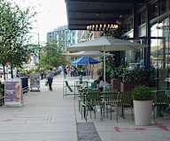 A sidewalk with tables and chairs and people walking.