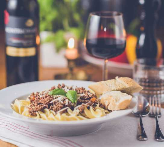 A plate of pasta with a glass of red wine and a bottle of wine in the background.