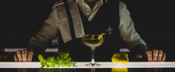 A person in a vest is leaning over a table with a cocktail glass, a sprig of mint, and a lemon wedge on it. at The Lady, South Carolina