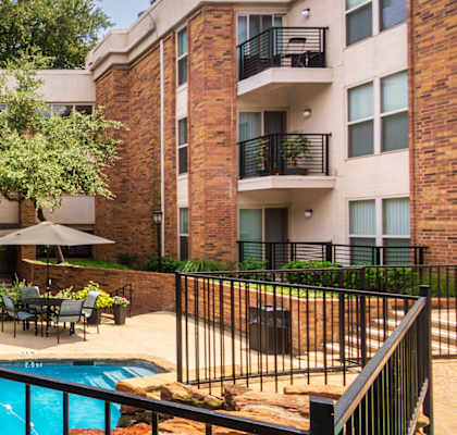 Swimming pool with a waterfall surrounded by lounge chairs and a couple of tables with chairs and umbrellas at Cambridge Court Apartments in the Lake Highlands neighborhood of Dallas, TX.