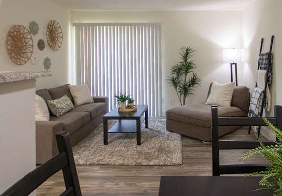 View of the living room with a couch, chaise lounge and a potted plant from the dining room of a 515 square foot 1 bedroom, 1 bath apartment home at Canyon Creek Apartments in Dallas Midtown neighborhood of Dallas, TX.