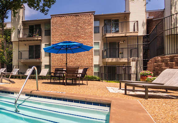 A swimming pool surrounded by chairs and an umbrella in with apartments in the background at Canyon Creek in the Midtown neighborhood of Dallas, TX.