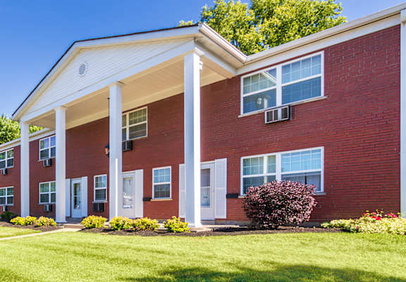 Brick apartment building exterior with sidewalk, lawn, and landscaped beds at Lake of the Woods Apartments