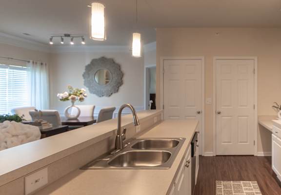 View down the Islander kitchen past the sink and countertops, with a refrigerator, white cabinets, pendant lights, and the adjoining living space beyond.