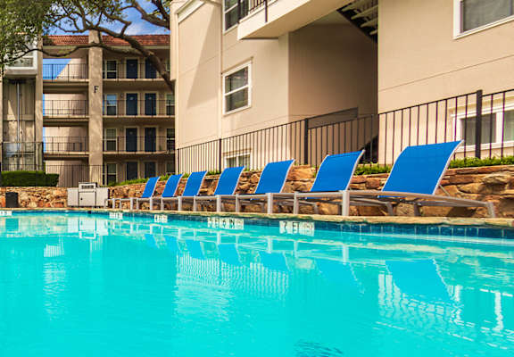 A pool area with lounge chairs, a patio table and chairs with a blue umbrella and stainless steel BBQ in front of apartment buildings at Princeton Court Apartments in the Vickery Midtown neighborhood of Dallas, TX.