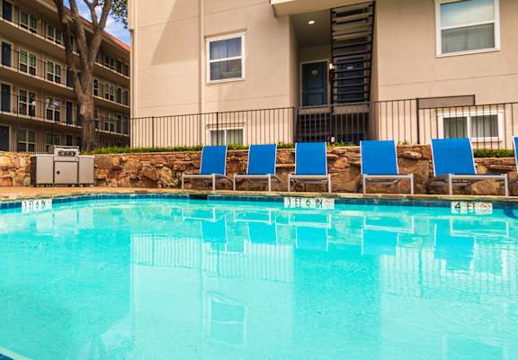 A pool area with lounge chairs, a patio table and chairs with a blue umbrella and stainless steel BBQ in front of apartment buildings at Princeton Court Apartments in the Vickery Midtown neighborhood of Dallas, TX.