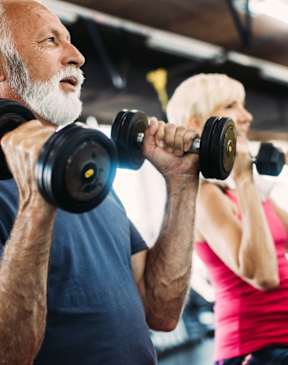 An older man and a younger woman are working out with dumbbells in a gym.