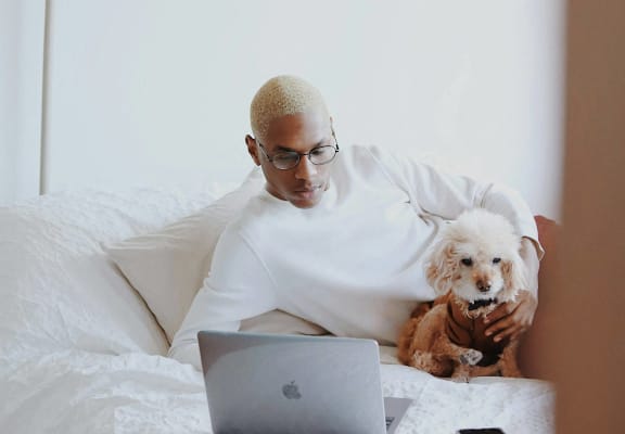 a man sitting on a bed with a dog looking at a laptop