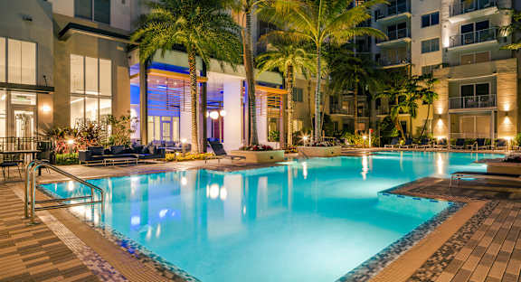 A swimming pool surrounded by palm trees and lounge chairs at night.