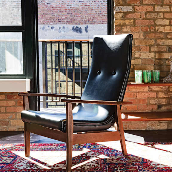 A black leather chair with wooden legs sits on a rug in front of a brick wall.