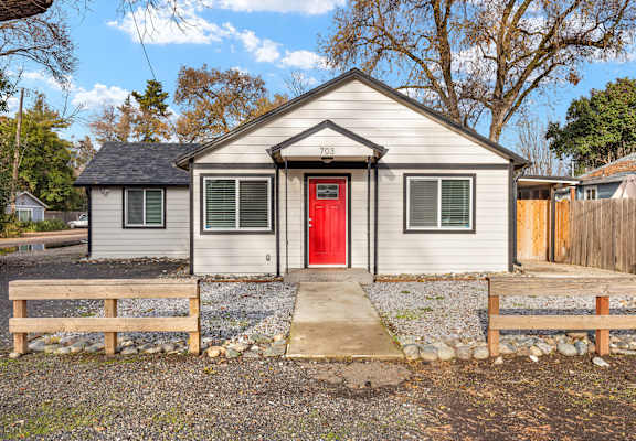 A small house with a red door is surrounded by a gravel area and wooden fences.