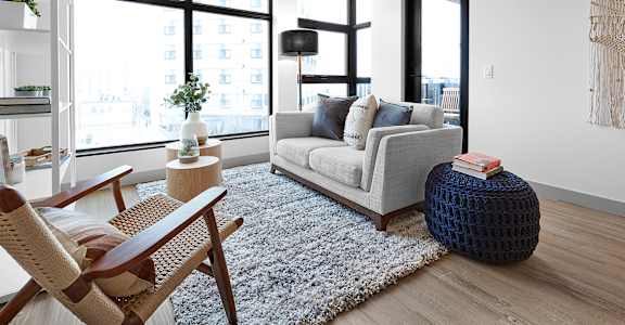 A living room with a grey couch, a wooden chair, a white rug, and a large window.at Raymond Block, Edmonton Alberta T6E 3R7