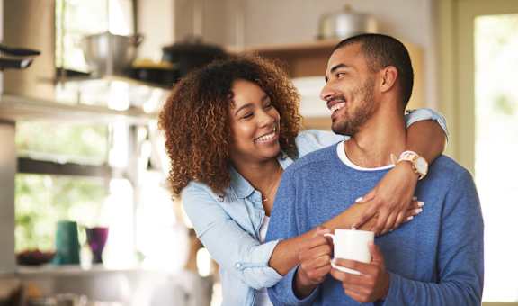 A man and woman are standing in a kitchen, the woman is holding a mug and they are both smiling.