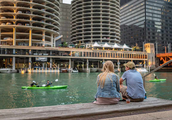 Two people sitting on a dock watching kayakers in the water.