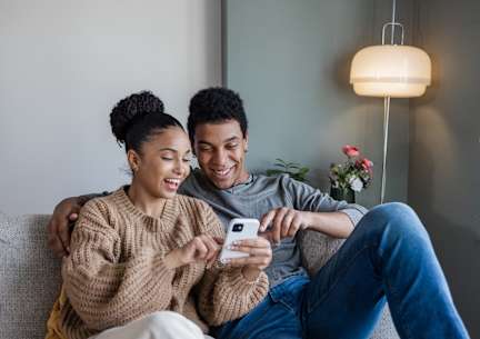 A couple is sitting on a couch, with the woman holding a smartphone and both of them smiling.