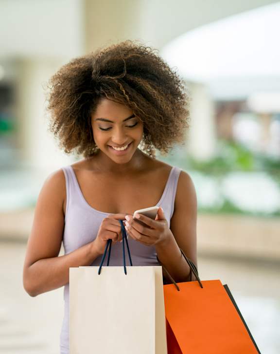 A woman with a white tank top is holding shopping bags and looking at her phone.