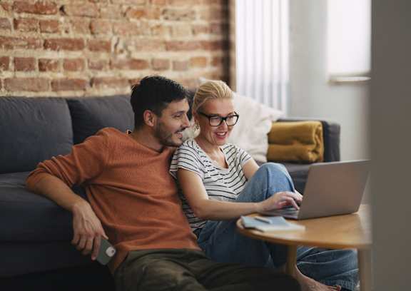 A man and a woman are sitting on a couch with a laptop on a table in front of them.