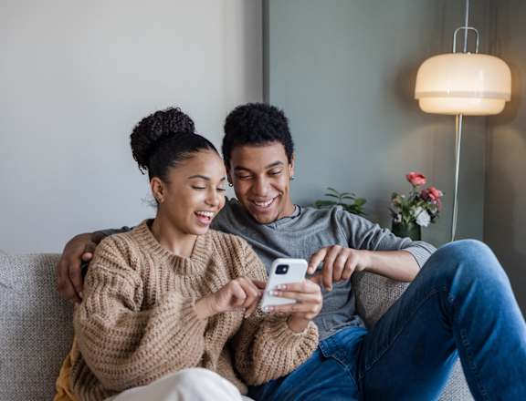 A couple is sitting on a couch, with the woman holding a smartphone and both of them smiling.