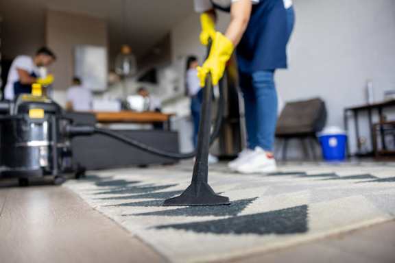 A person in a blue shirt and yellow gloves is vacuuming a rug in a room with other people in the background.