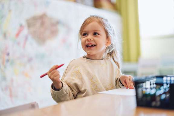 A young girl is smiling and holding a red pen while writing on a piece of paper. at The Woodlands Apartments, Milton Keynes